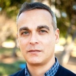 Man wearing blue collared shirt and sweater against natural backdrop